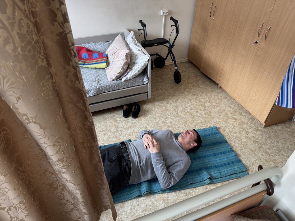 Man lying on a blue mat in a room with a walker, a couch, and a neatly made bed, suggesting a focus on accessibility and comfort in home care.