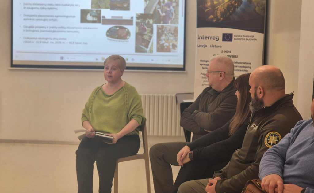 Panel discussion featuring a woman in a green top and three seated participants, with a presentation screen displaying images and information in the background. Panel discussion featuring a woman in a green top and three seated participants, with a presentation screen displaying images and information in the background.