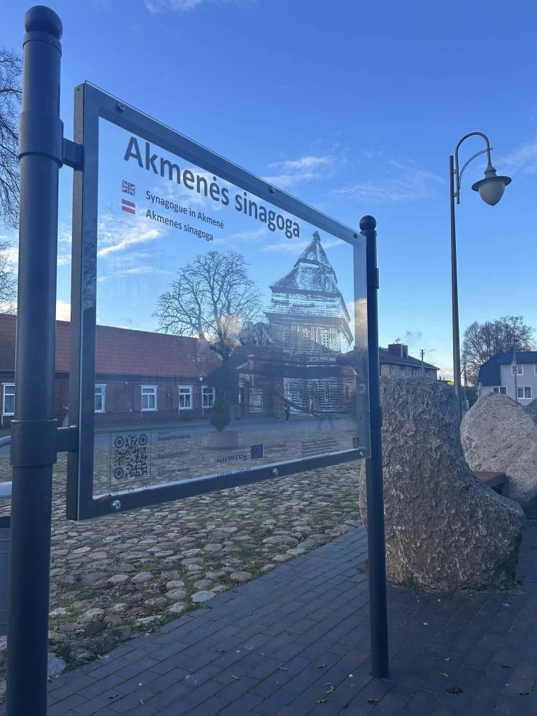 Signpost for Akmenes stacija, featuring historical information and a view of nearby buildings under a clear blue sky. Signpost for Akmenes stacija, featuring historical information and a view of nearby buildings under a clear blue sky.