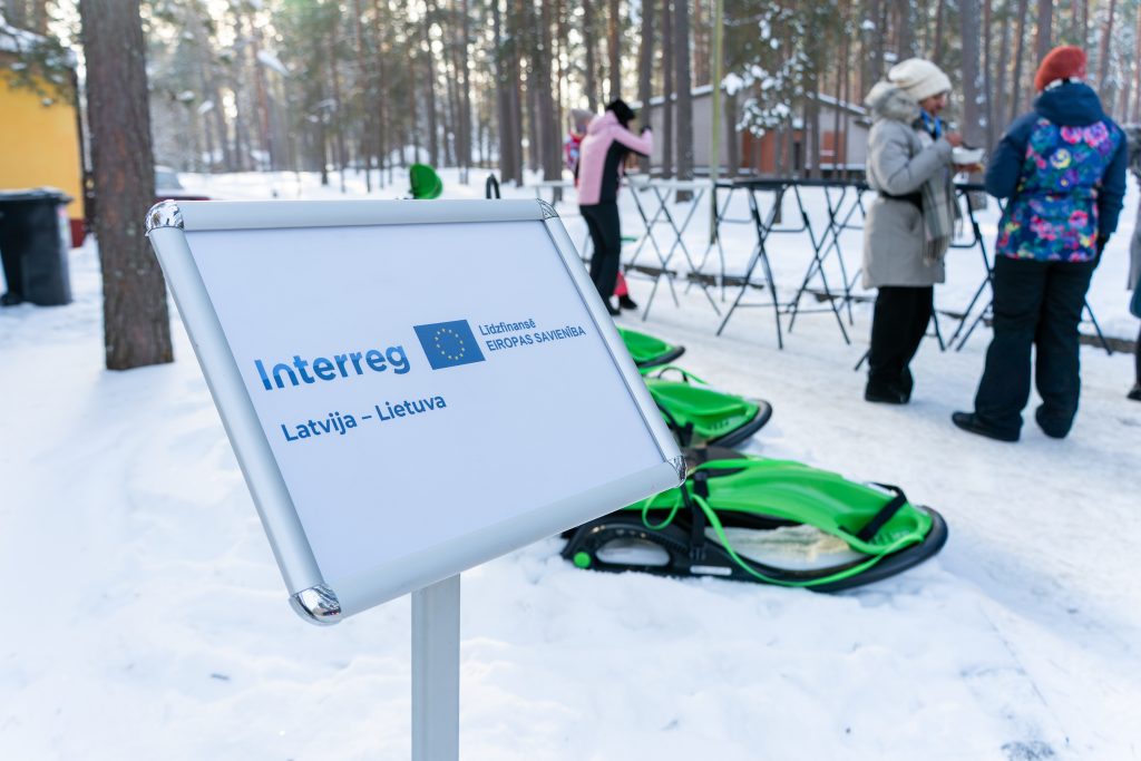 Signage for the Interreg project in a snowy landscape, with people preparing for winter activities and green sleds nearby.