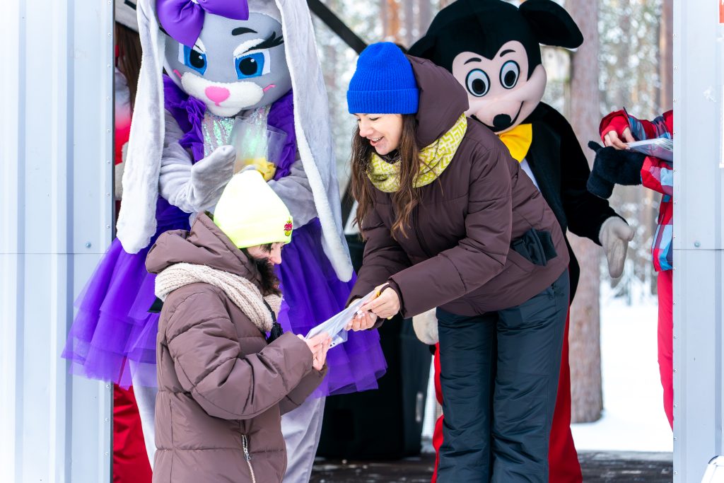 A woman in a brown jacket hands a gift to a girl in a yellow hat, surrounded by costumed characters including a purple bunny and a classic cartoon mouse, at a festive outdoor event.