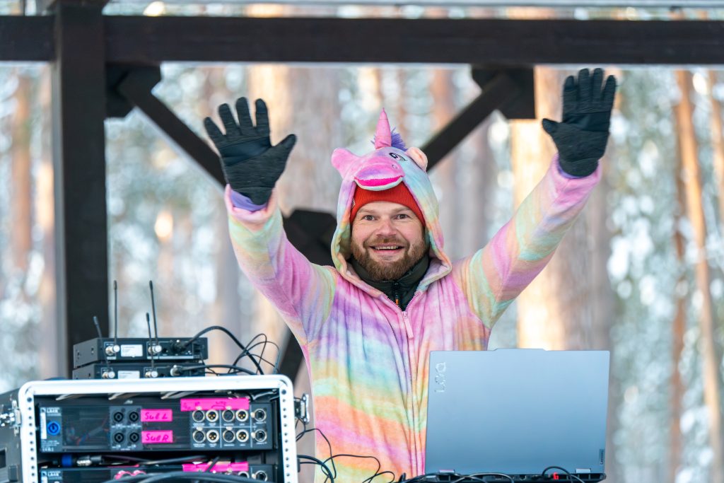 DJ in a colorful unicorn onesie performing at an outdoor music festival, surrounded by trees and a sound setup.