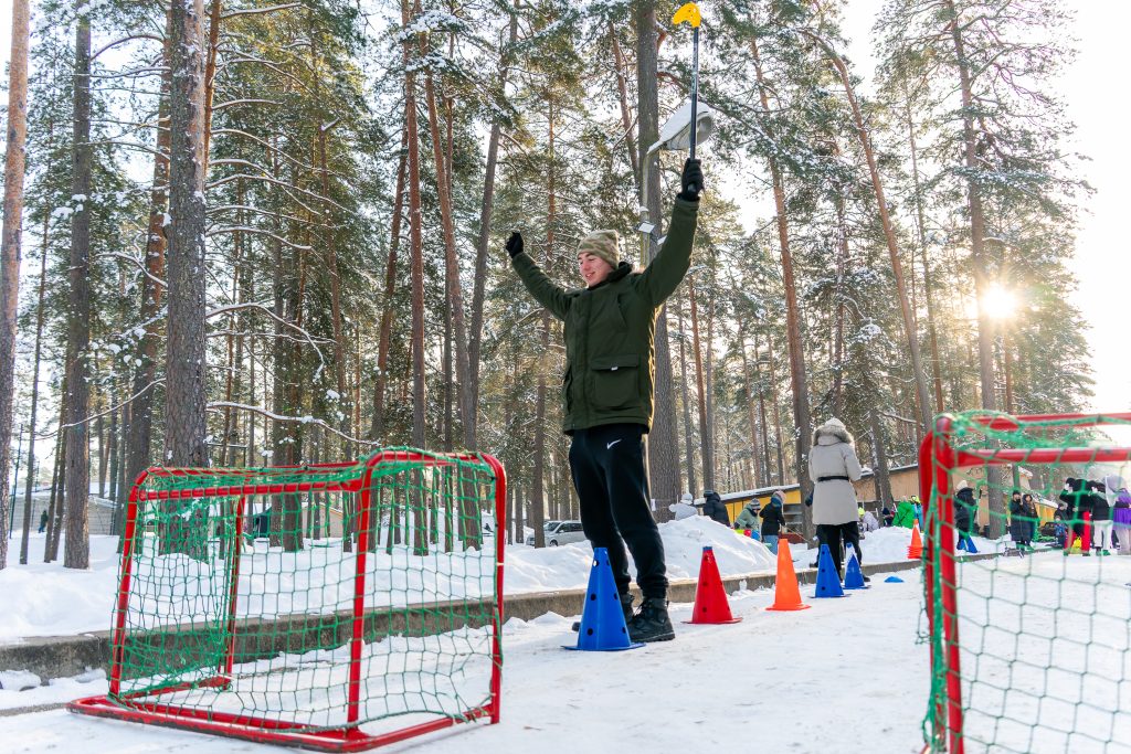 Person celebrating in a snowy forest setting, raising arms in excitement while standing on a blue cone, surrounded by colorful traffic cones and small goals, with other people in the background.