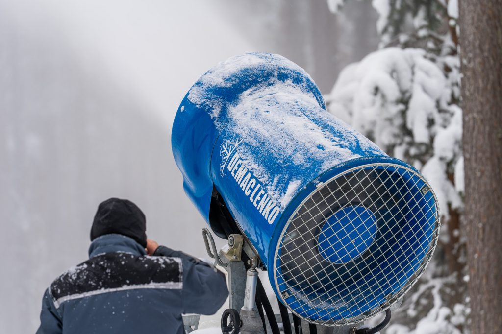 Snowmaking machine in operation, producing artificial snow in a winter landscape. A person adjusts the equipment in a snowy environment, showcasing winter sports preparation.