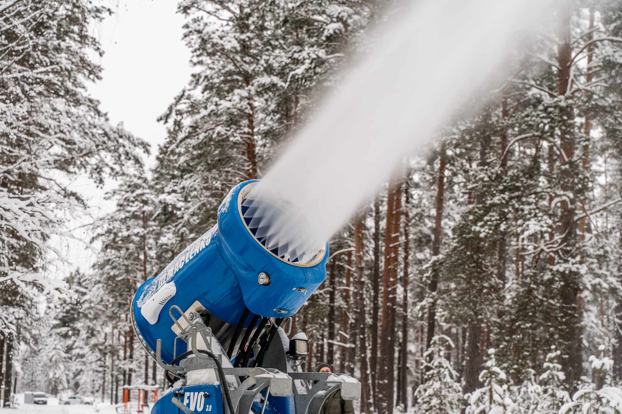 Snow cannon producing artificial snow in a snowy forest, surrounded by tall pine trees.
