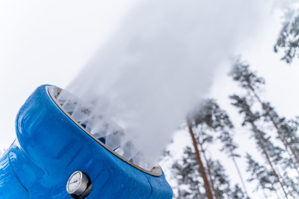 Blue snow cannon producing artificial snow in a winter landscape with trees in the background.