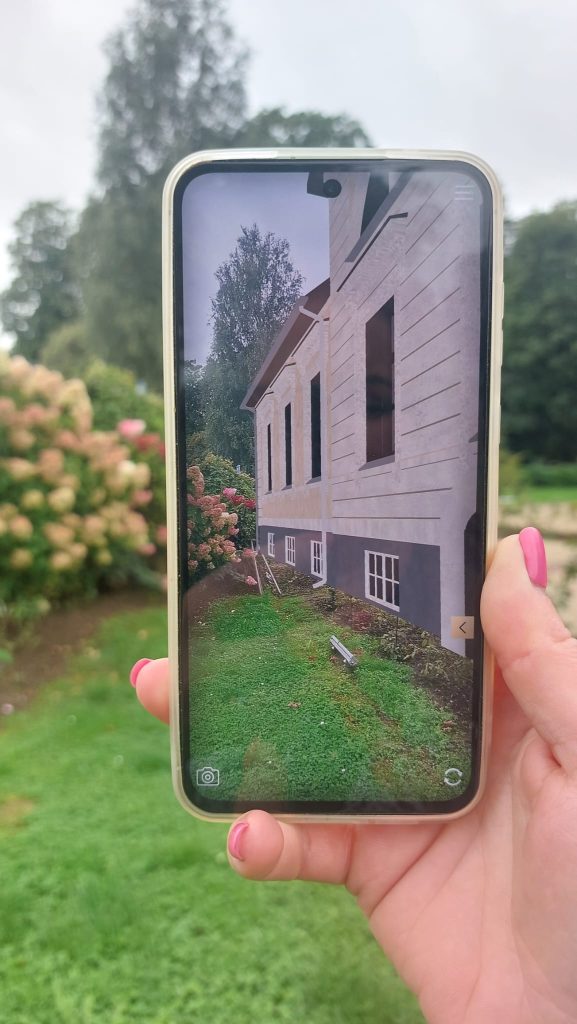 A person holding a smartphone displaying an image of a house's exterior, showcasing a landscaped garden with hydrangeas and a clear sky in the background. The image highlights architectural details like windows and siding. A person holding a smartphone displaying an image of a house's exterior, showcasing a landscaped garden with hydrangeas and a clear sky in the background. The image highlights architectural details like windows and siding.