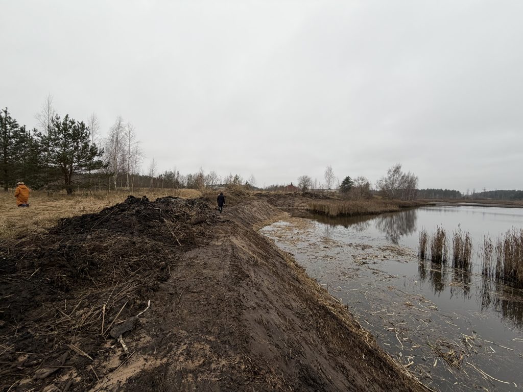 Two people walking along a muddy embankment beside a calm river with sparse vegetation and overcast skies, showcasing a natural landscape affected by recent weather conditions. Two people walking along a muddy embankment beside a calm river with sparse vegetation and overcast skies, showcasing a natural landscape affected by recent weather conditions.