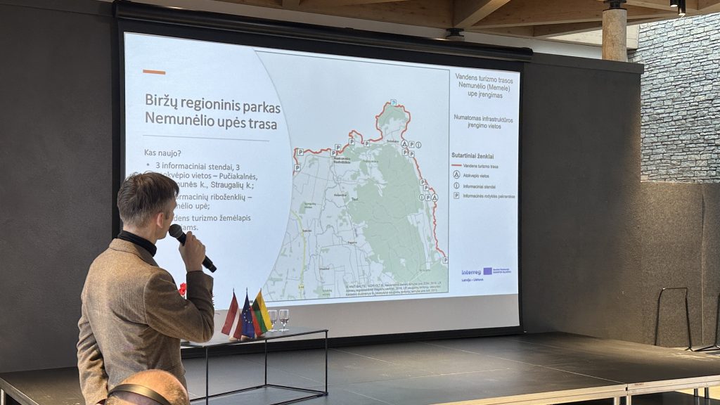 A presenter speaking at a conference, with a map displayed on a screen showing community development areas, alongside flags of various countries in the foreground. A presenter speaking at a conference, with a map displayed on a screen showing community development areas, alongside flags of various countries in the foreground.