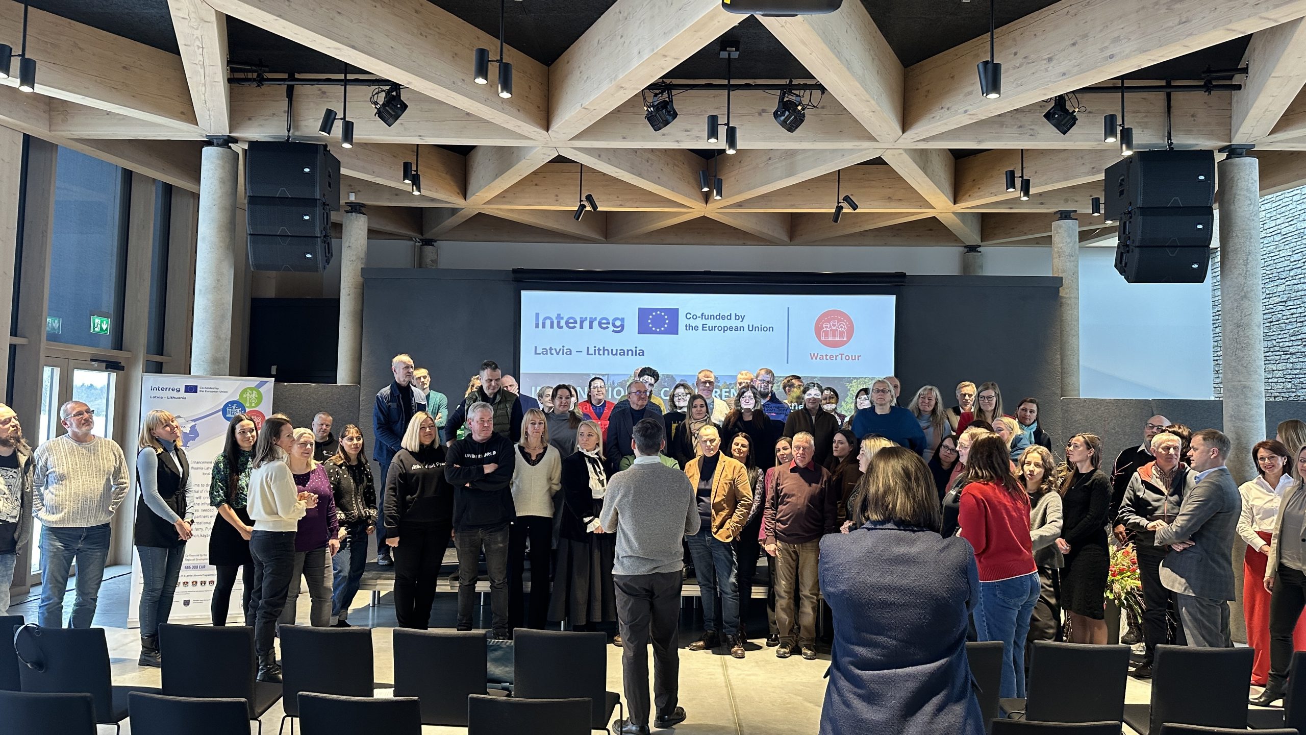 Group photo of participants at a conference, featuring diverse attendees standing together in a modern venue with a large screen displaying logos for the European Union and a local organization. Group photo of participants at a conference, featuring diverse attendees standing together in a modern venue with a large screen displaying logos for the European Union and a local organization.