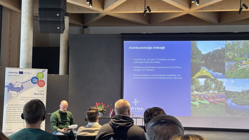 A speaker presenting in front of an audience during a seminar, with a blue presentation slide visible in the background. The setting features modern architecture and floral arrangements, highlighting an engaging educational environment. A speaker presenting in front of an audience during a seminar, with a blue presentation slide visible in the background. The setting features modern architecture and floral arrangements, highlighting an engaging educational environment.