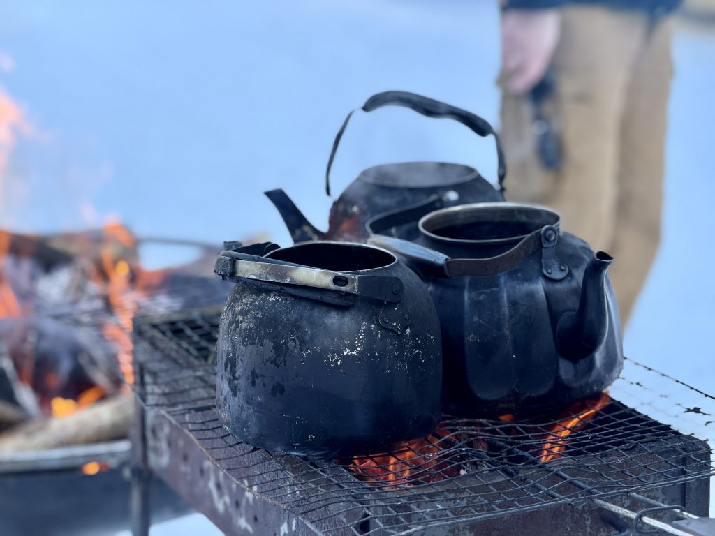 Black metal kettles resting on a grill over an open fire, with a blurred figure in the background, set against a snowy landscape.