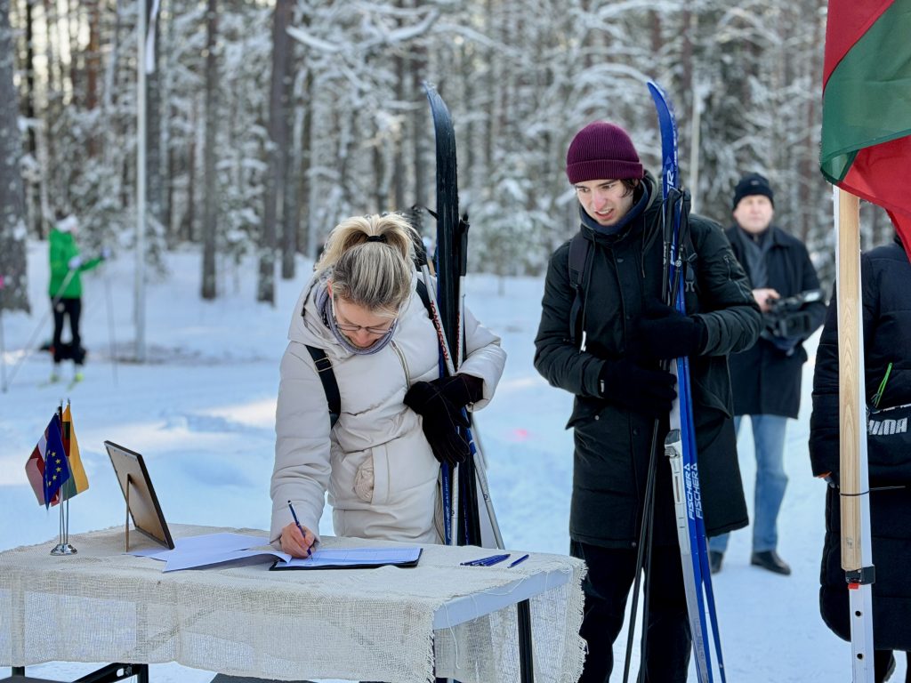Two people preparing for cross-country skiing in a snowy forest, with one individual signing in at a registration table while the other holds ski poles.