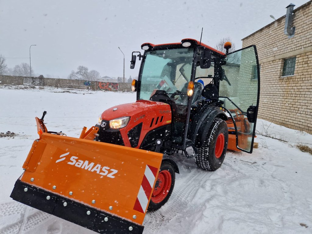 Orange tractor with a snow plow attachment parked on a snowy surface, showing a snowy landscape and a brick building in the background. The tractor is equipped with a front loader and has its door open, indicating it is ready for snow removal tasks.