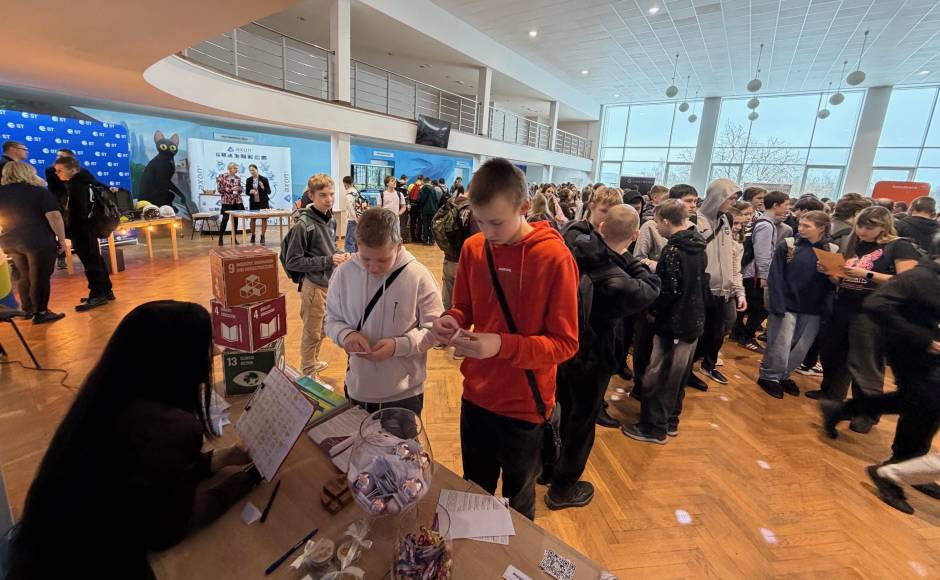 A crowded event space with attendees lining up at a booth, featuring two young men in the foreground, one wearing a red hoodie and the other in a white shirt, both holding mobile devices. A crowded event space with attendees lining up at a booth, featuring two young men in the foreground, one wearing a red hoodie and the other in a white shirt, both holding mobile devices.