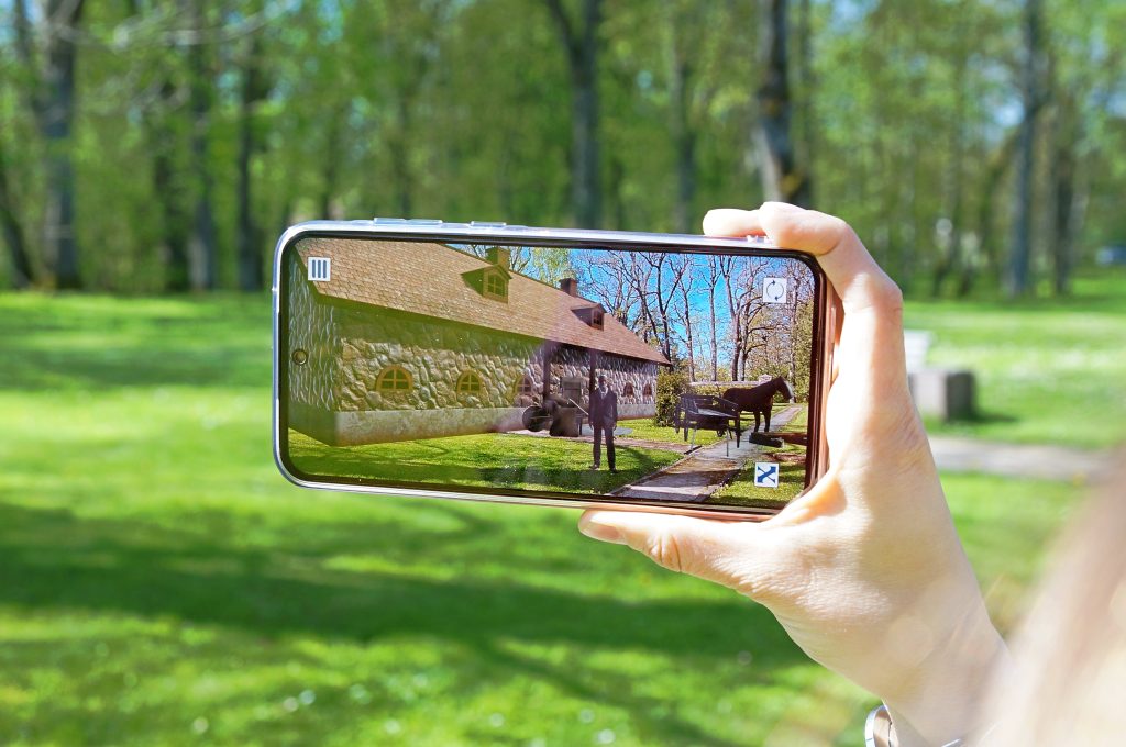 Person holding a smartphone displaying a picturesque view of a stone house and green landscape, surrounded by trees in bright sunlight. Person holding a smartphone displaying a picturesque view of a stone house and green landscape, surrounded by trees in bright sunlight.