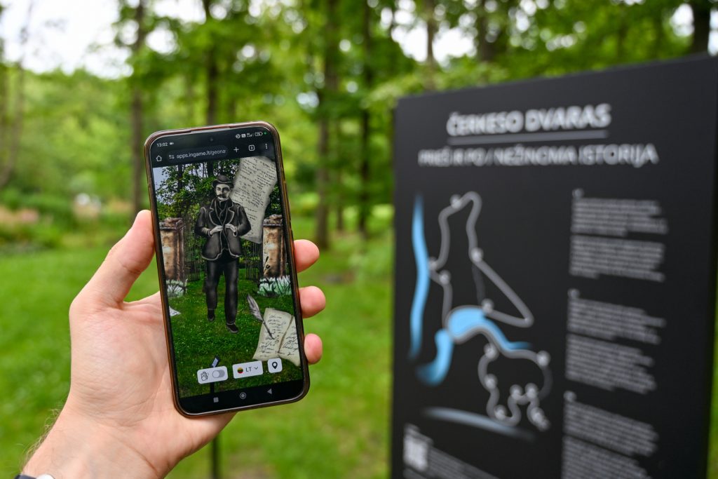 Person holding a smartphone displaying an augmented reality experience featuring a bear, with an informational sign in a forested area. Person holding a smartphone displaying an augmented reality experience featuring a bear, with an informational sign in a forested area.