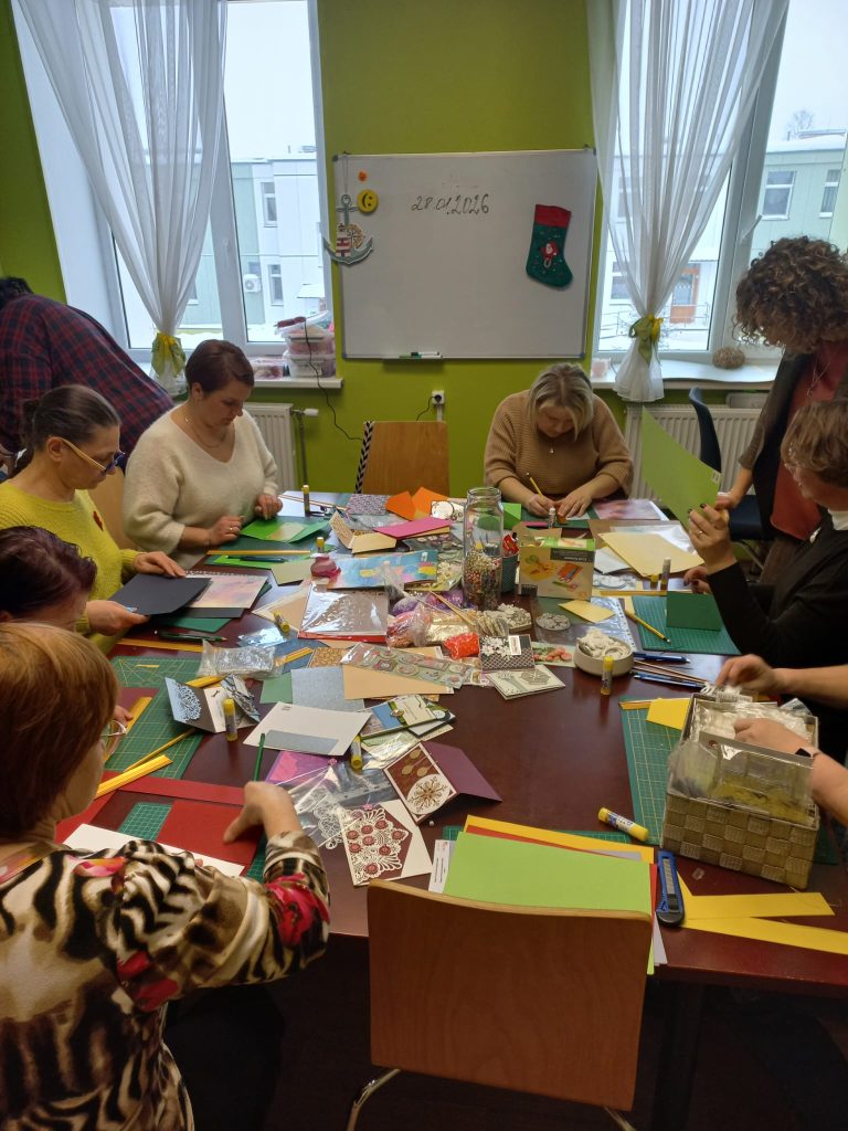 Group of people engaged in a crafting workshop, working with colorful paper and various materials on a table, surrounded by a bright and inviting classroom environment.
