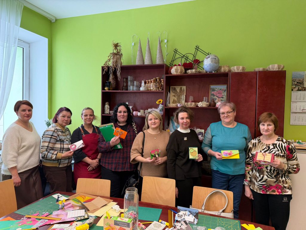 Group of women participating in a crafting workshop, showcasing their handmade projects and materials in a brightly colored room with shelves of decorative items.