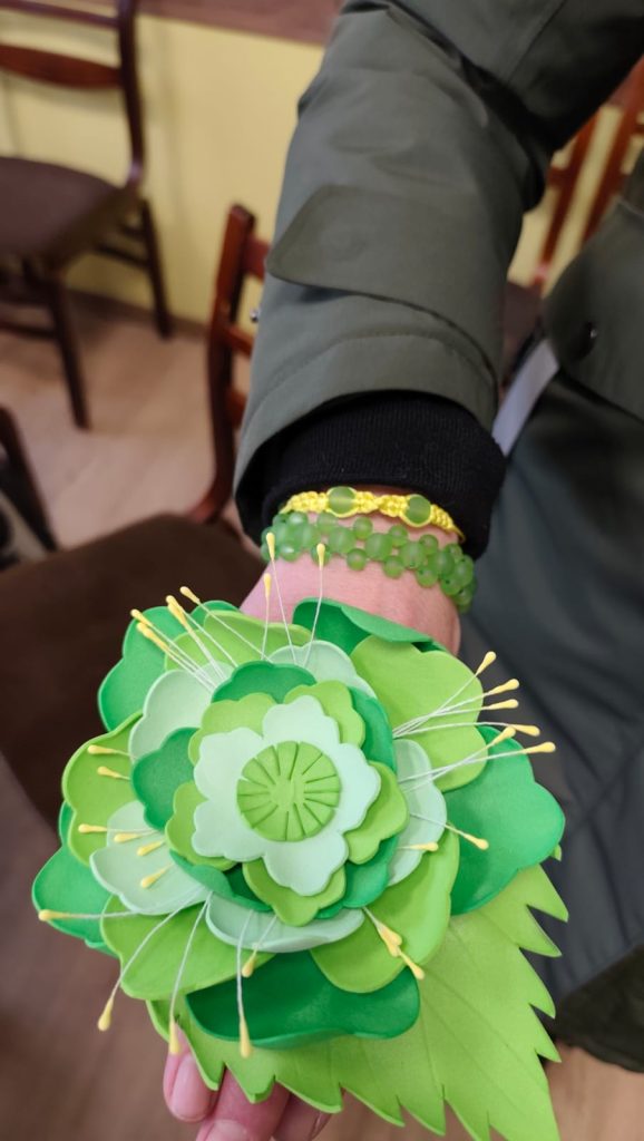 Close-up of a hand wearing a vibrant green flower wrist corsage, adorned with pins and beads, showcasing intricate paper crafting. The background features a blurred indoor setting.