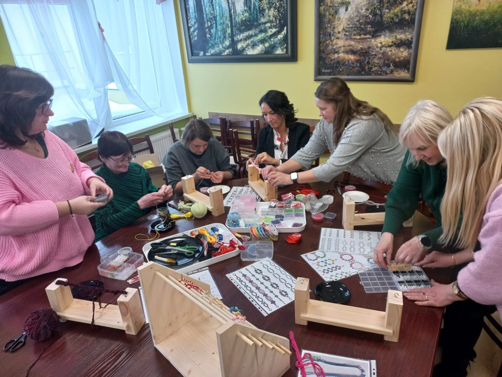 Group of people engaged in a crafting workshop, working on various projects with tools and materials on a table. The setting features a bright room with artwork on the walls, highlighting creativity and collaboration.