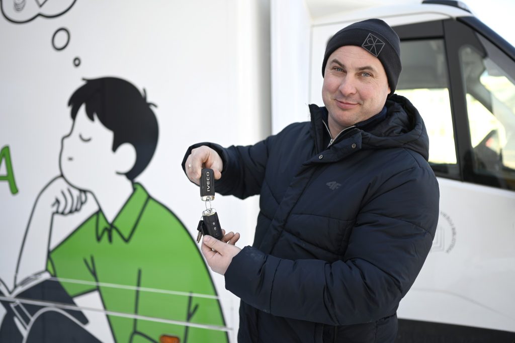 Man in a black jacket and beanie holding car keys, standing next to a colorful mural of a boy in a green shirt, showcasing urban art and community engagement.