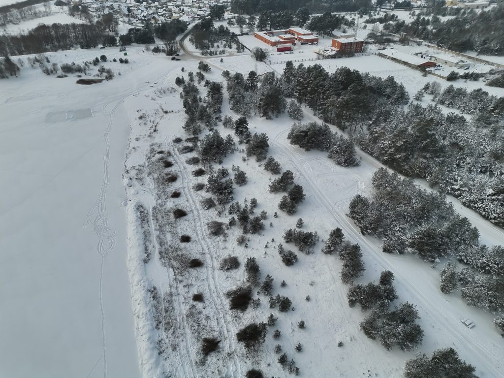 Aerial view of a snowy landscape featuring a frozen lake, tree-lined shoreline, and distant buildings, showcasing a serene winter scene. Aerial view of a snowy landscape featuring a frozen lake, tree-lined shoreline, and distant buildings, showcasing a serene winter scene.