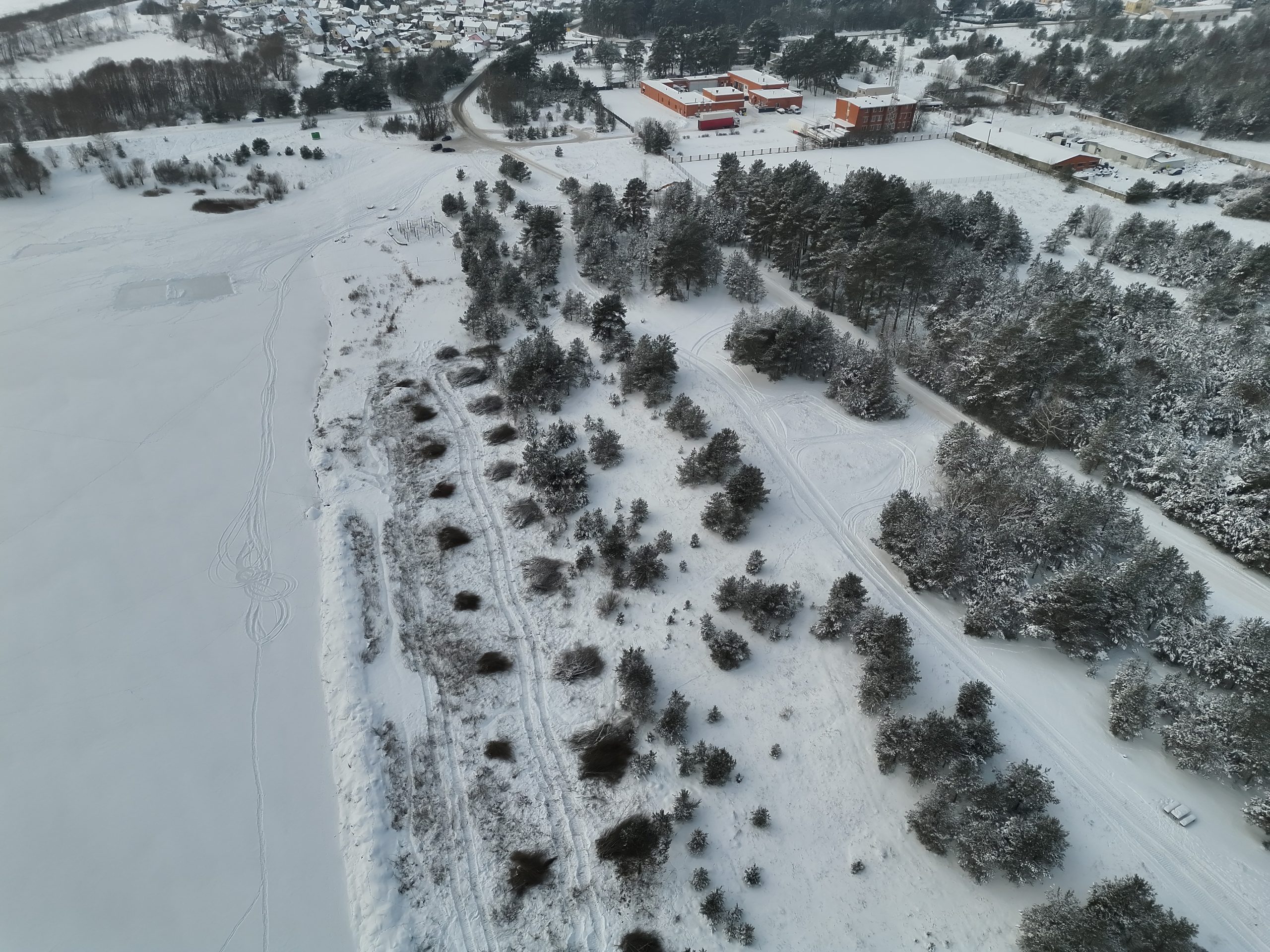 Aerial view of a snowy landscape featuring a frozen lake, tree-lined shoreline, and distant buildings, showcasing a serene winter scene.