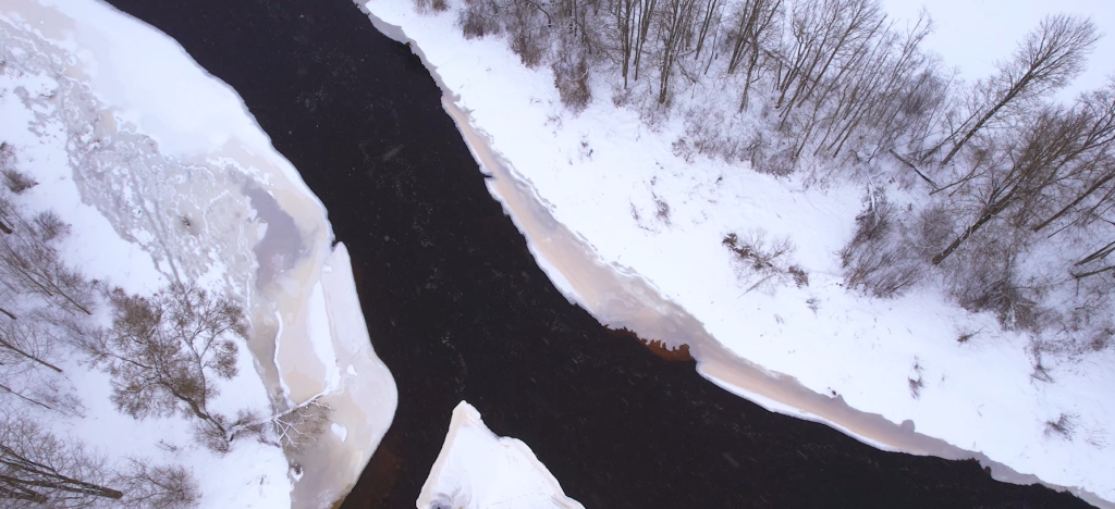 Aerial view of a winding river surrounded by snow-covered banks and trees, showcasing a contrast between dark water and white ice formations in a winter landscape.