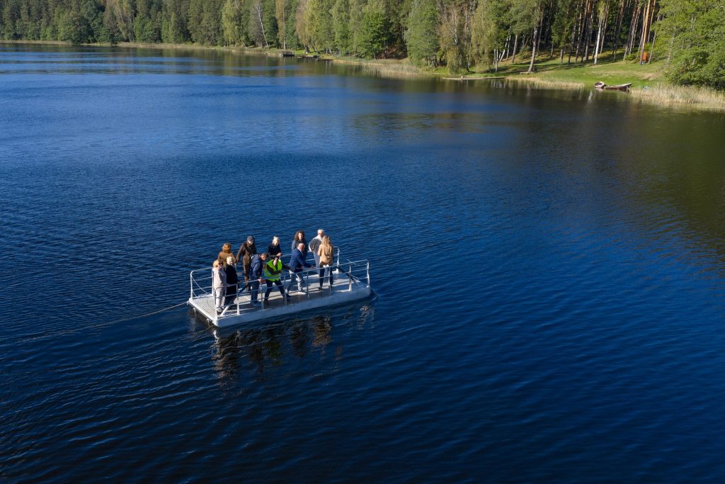 Group of people standing on a floating platform in a serene lake surrounded by lush green trees, showcasing outdoor recreational activities and nature exploration.