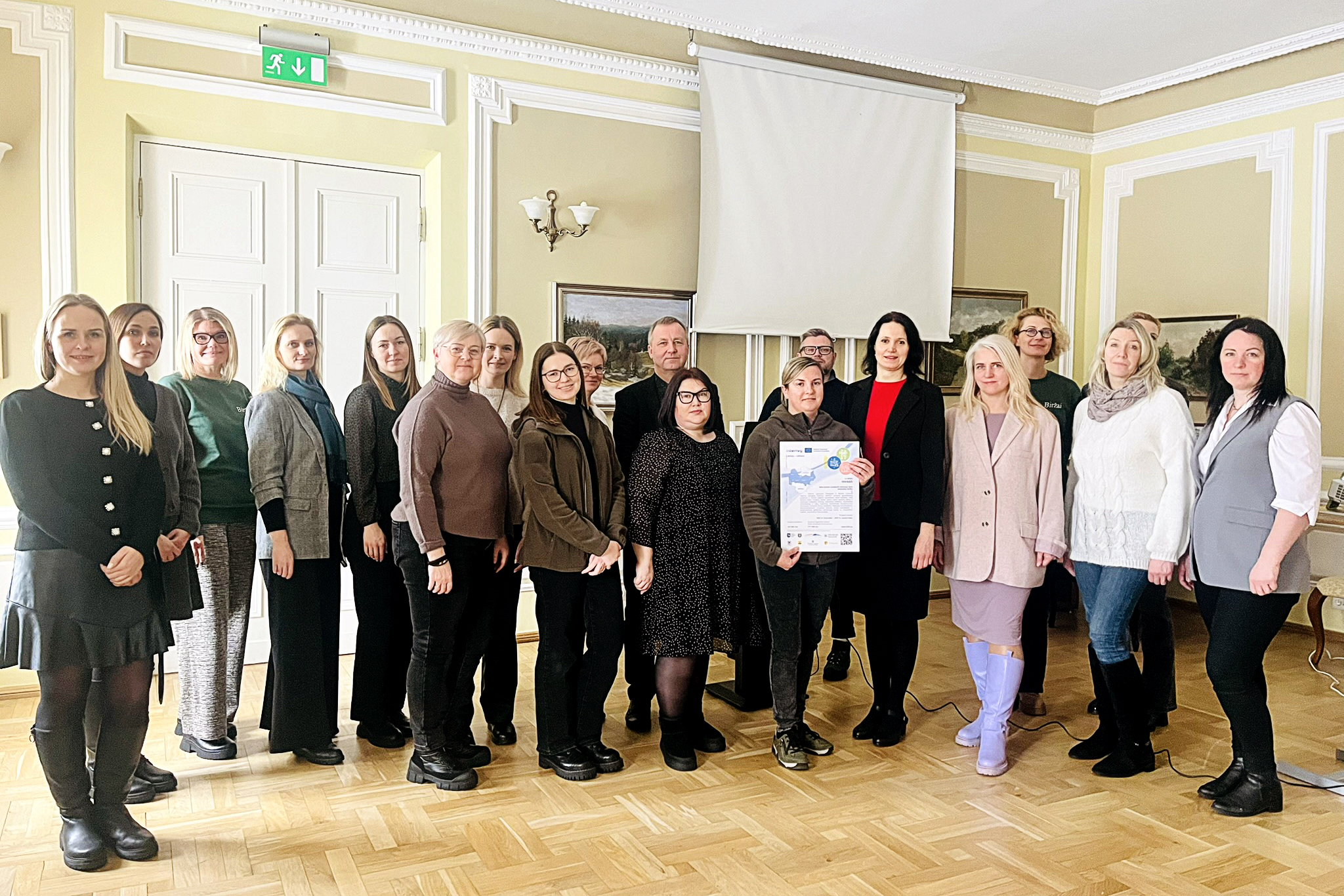 Group photo of a diverse team gathered for a workshop or meeting, standing in a well-lit room with a projector screen and artwork in the background. Group photo of a diverse team gathered for a workshop or meeting, standing in a well-lit room with a projector screen and artwork in the background.