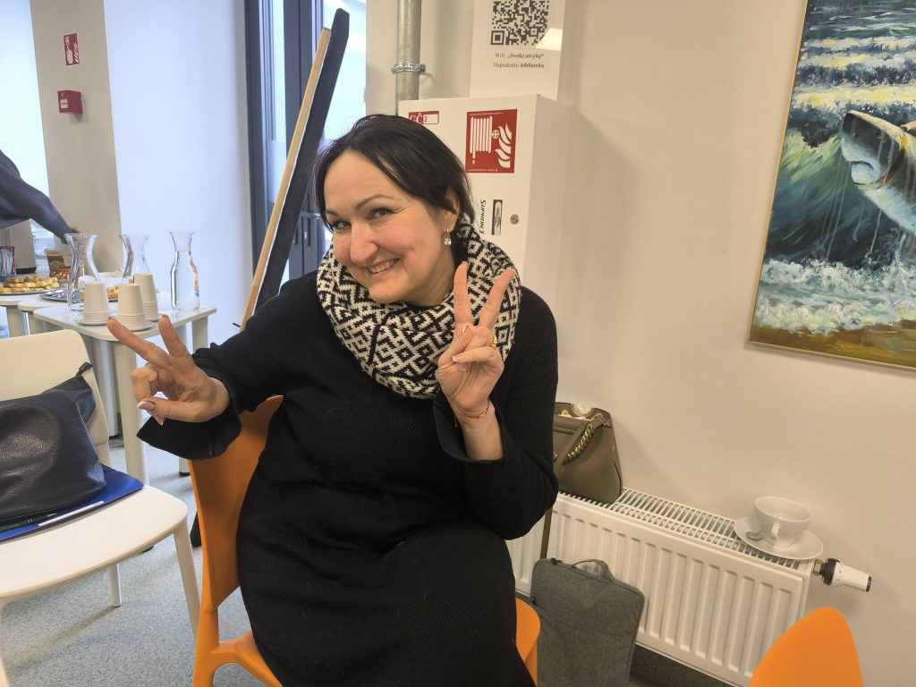 Woman smiling and making a peace sign while seated in a modern interior setting, with refreshments and artwork in the background. Ideal for topics related to social gatherings, events, or community engagement.