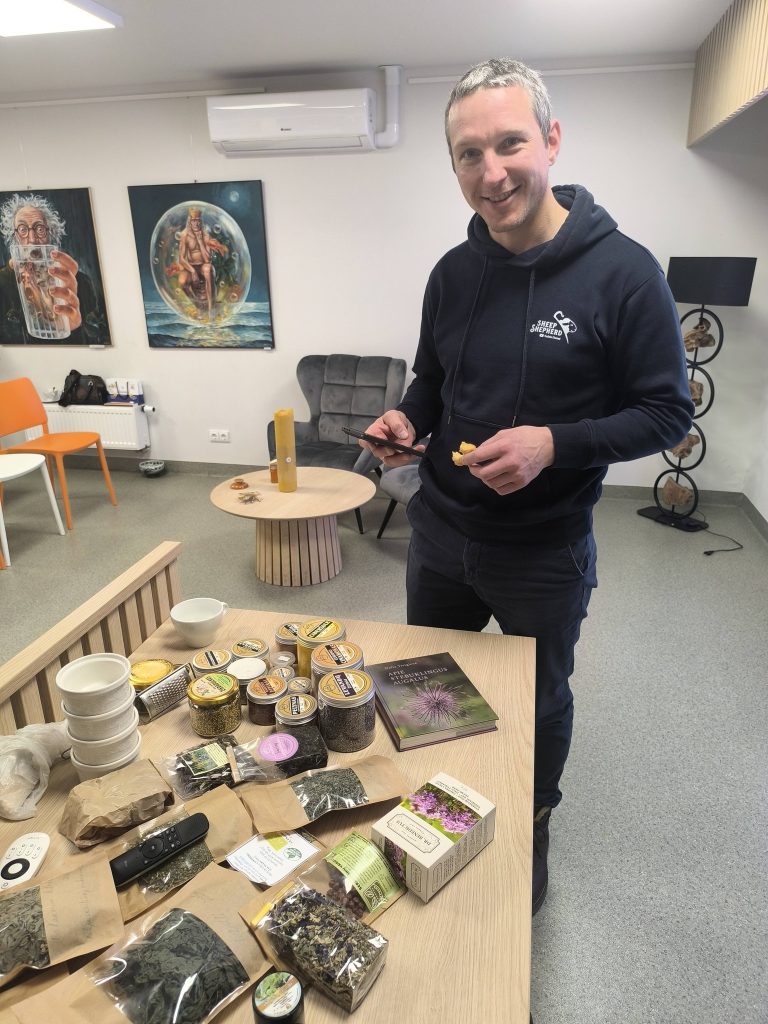 A man standing in a modern room with artwork on the walls, holding a piece of food in one hand and a phone in the other, surrounded by various jars and packages of herbal teas and ingredients on a table.