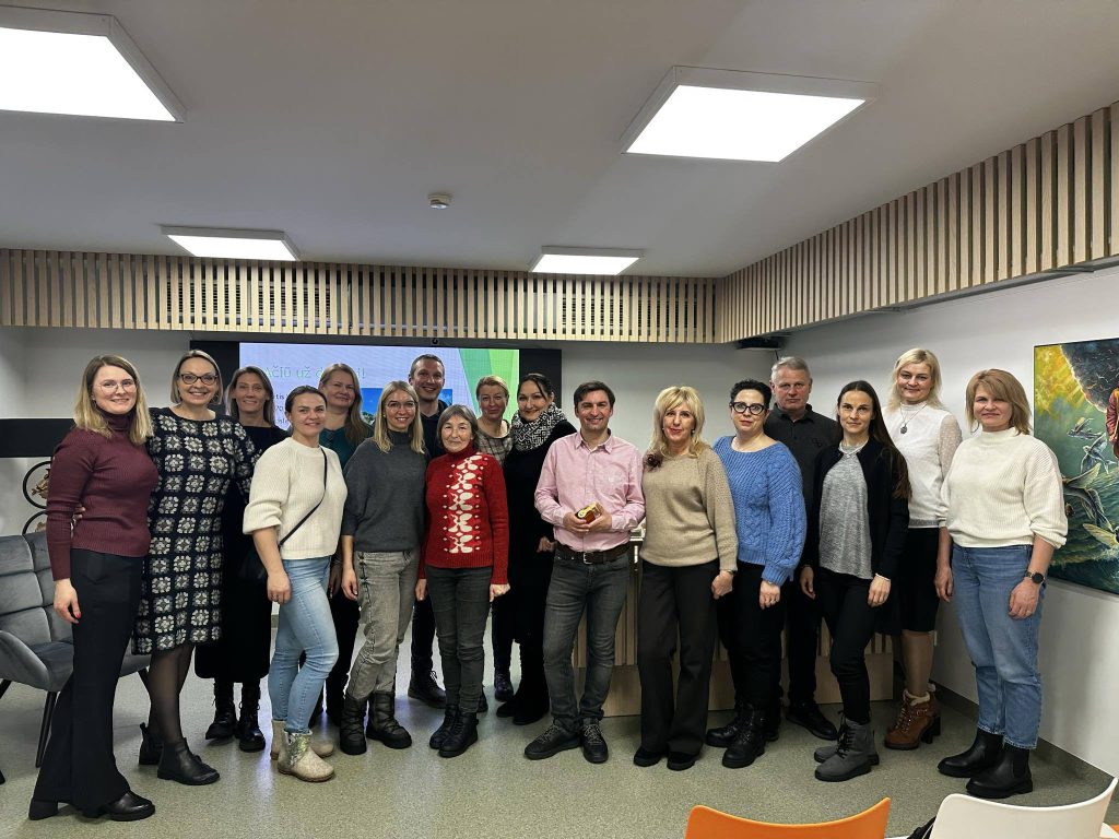 Group photo of a diverse team gathered in a modern office space, featuring men and women of various ages. The setting includes a presentation screen and artistic decor, highlighting collaboration and teamwork. Some participants are wearing festive attire, suggesting a celebratory occasion or event.