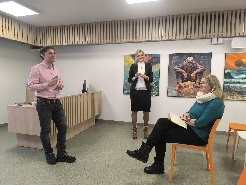 A business meeting in a modern office space featuring three professionals. A man in a pink shirt presents while a woman in a black blazer engages with him, and another woman sits with a notebook, observing. Colorful artwork adorns the walls, enhancing the creative atmosphere.