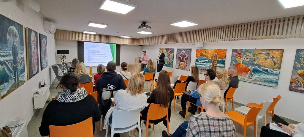 Audience attending a presentation in a modern conference room, featuring colorful artwork on the walls and a speaker engaging with attendees. The setting includes chairs in vibrant orange and neutral tones, with a projector screen displaying information.