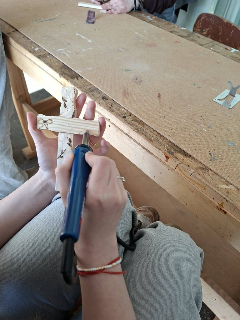 Person using a wood-burning tool to decorate a wooden cross in a workshop setting, with a focus on hands and the crafting process.