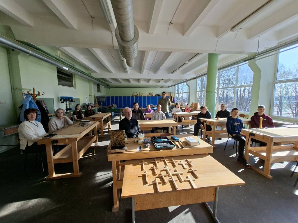 Students and instructors in a classroom with wooden work tables, engaged in a learning environment focused on hands-on projects. Natural light floods the space through large windows, highlighting the workshop's tools and materials.