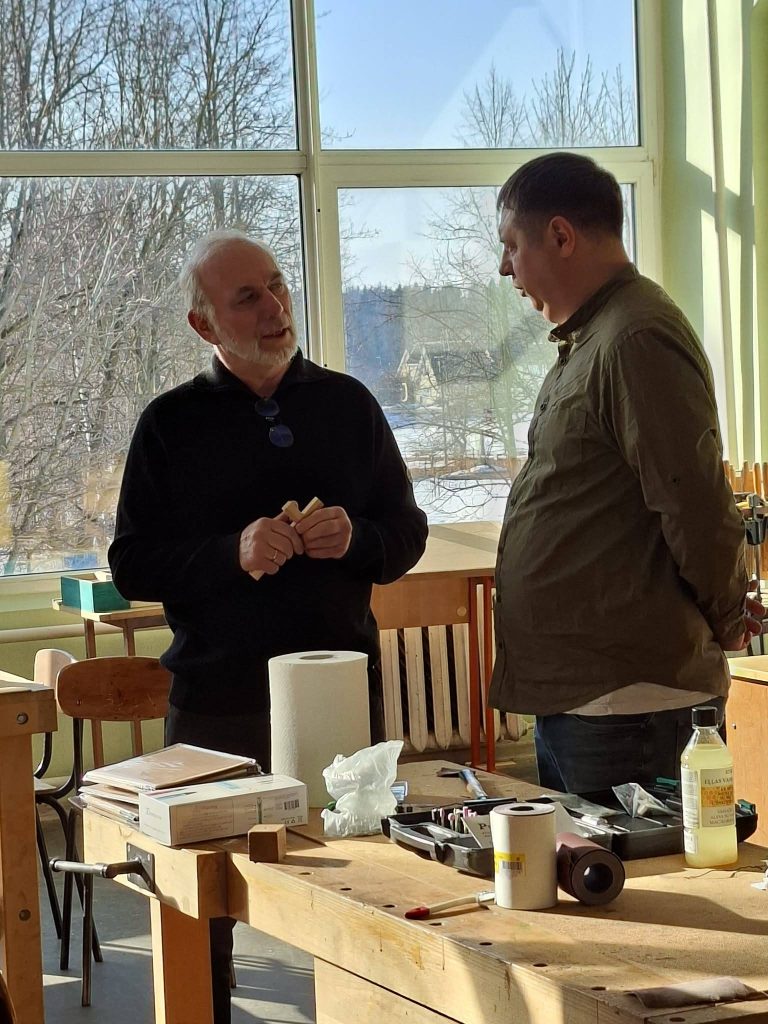 Two men engaged in a conversation in a well-lit workshop, discussing woodworking tools. The workspace features a wooden table with various tools, paper towels, and materials, while large windows provide a view of a snowy landscape outside.