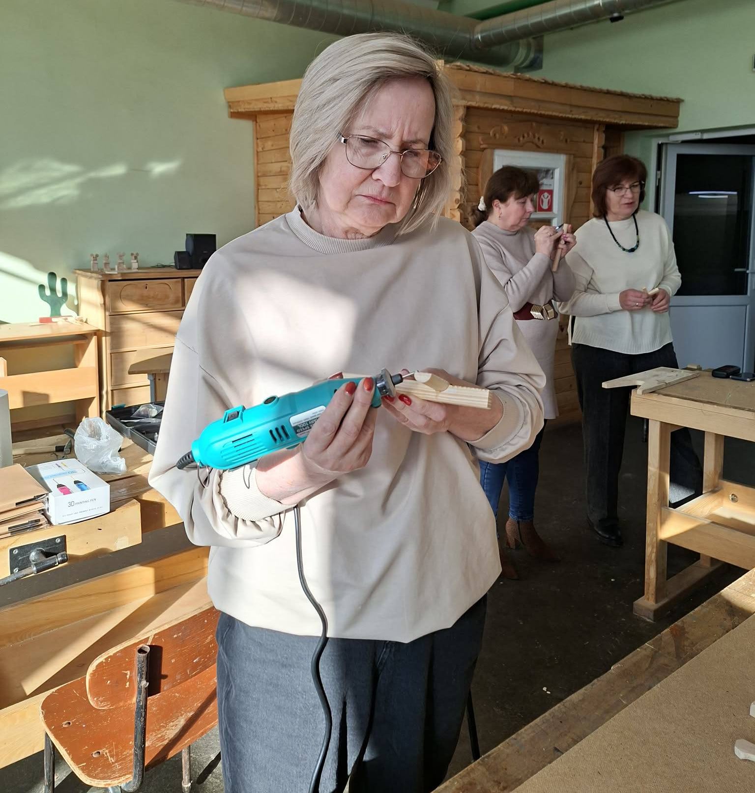 A woman using a power tool to craft wood in a workshop, with other participants engaged in activities in the background. The setting features wooden furniture and tools, highlighting a hands-on learning environment.