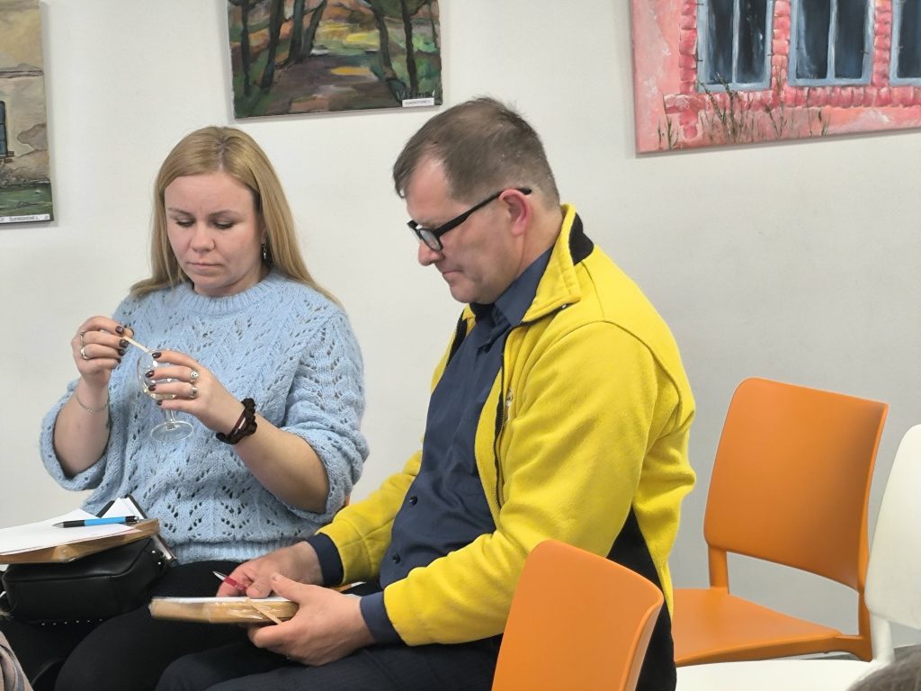 A woman in a light blue sweater examines a small glass object while a man in a yellow jacket and blue shirt sits next to her, holding a notebook. Both are engaged in a discussion, surrounded by colorful artwork on the walls and modern chairs.