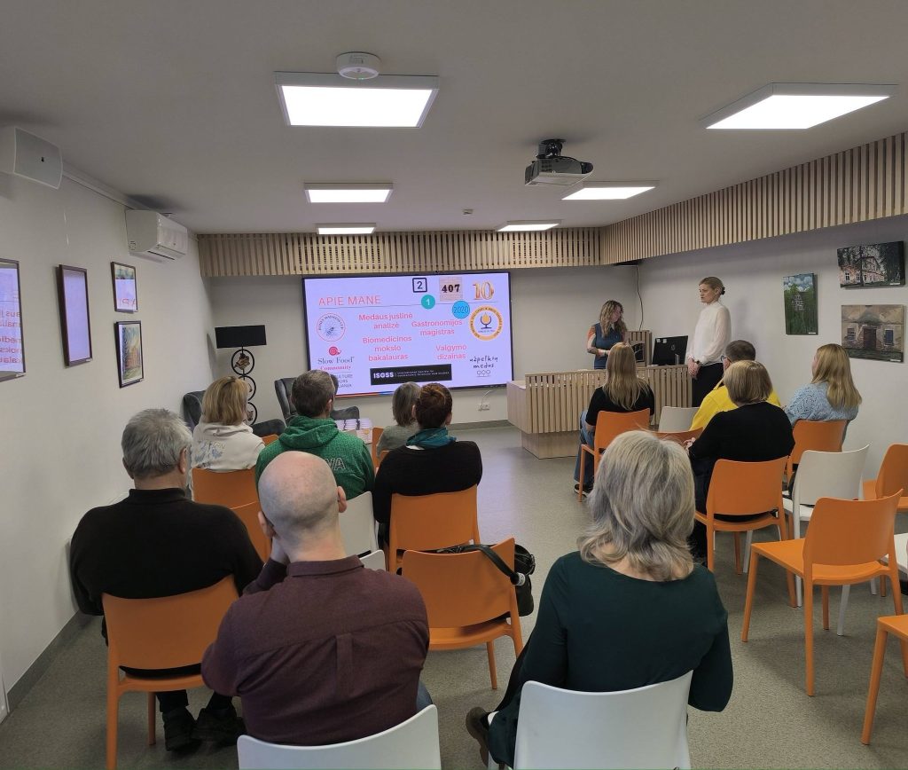 Audience attending a presentation in a modern seminar room, featuring two speakers discussing topics related to gastronomy and education, with a projector displaying key information on a screen. The room is equipped with comfortable seating and a contemporary design.