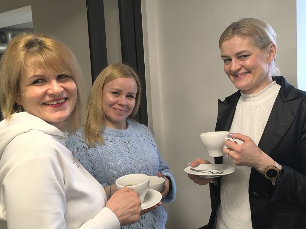 Three women smiling and holding cups of tea or coffee, standing together in a bright indoor setting, showcasing a friendly social gathering.