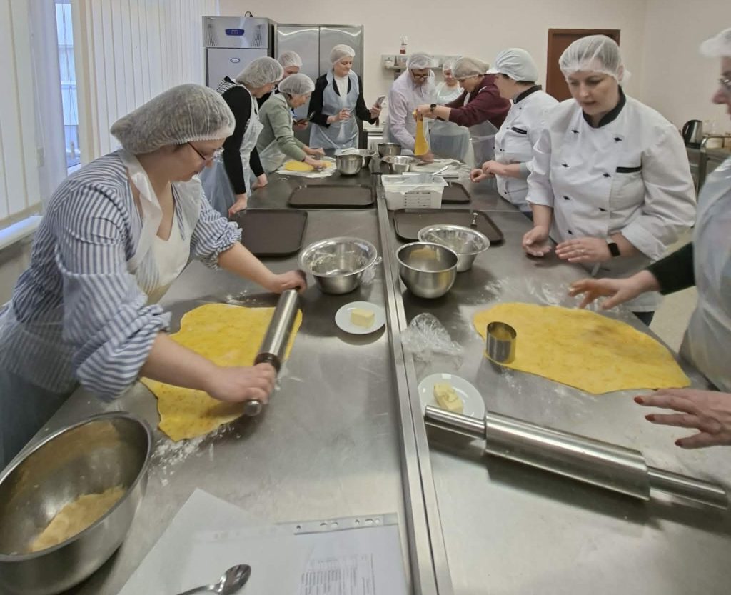 Group of culinary students in a professional kitchen, wearing hairnets and aprons, actively participating in a cooking class. They are rolling out dough and preparing ingredients on stainless steel counters, showcasing teamwork and culinary skills in a hands-on learning environment.
