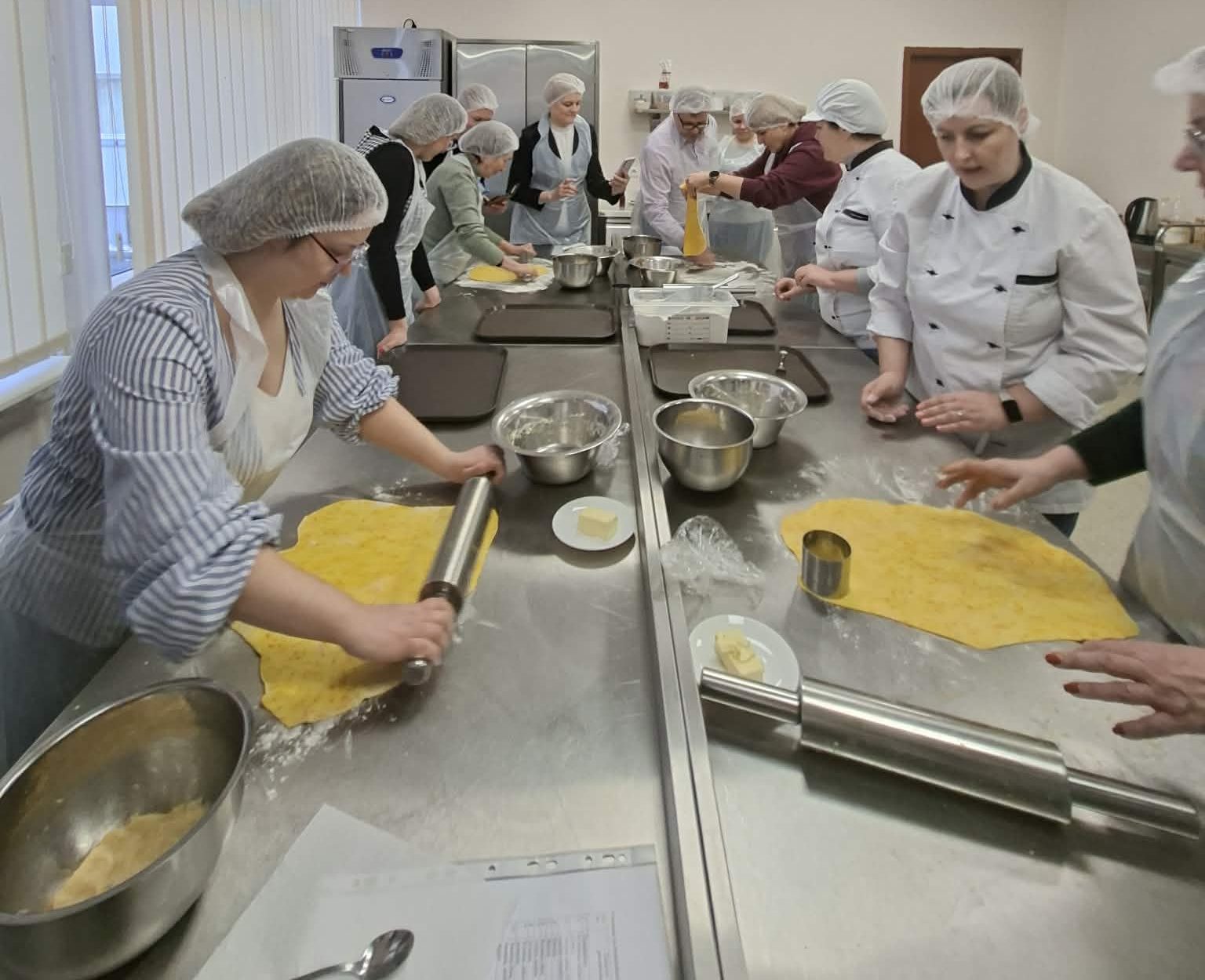 Group of culinary students in a professional kitchen, wearing hairnets and aprons, actively participating in a cooking class. They are rolling out dough and preparing ingredients on stainless steel counters, showcasing teamwork and culinary skills in a hands-on learning environment.
