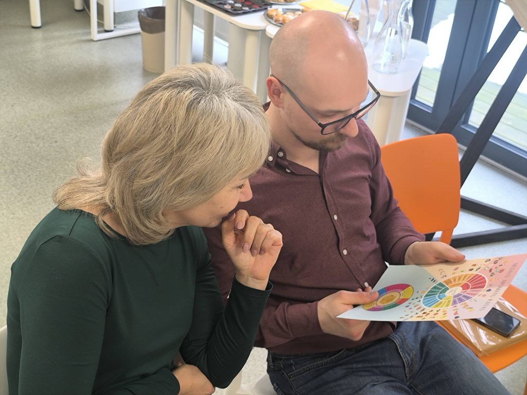 Two people engaged in a discussion while reviewing a colorful chart. The woman has shoulder-length blonde hair and wears a green top, while the man, who wears glasses and a maroon shirt, holds the chart. The setting appears to be a modern office with natural light and casual seating.