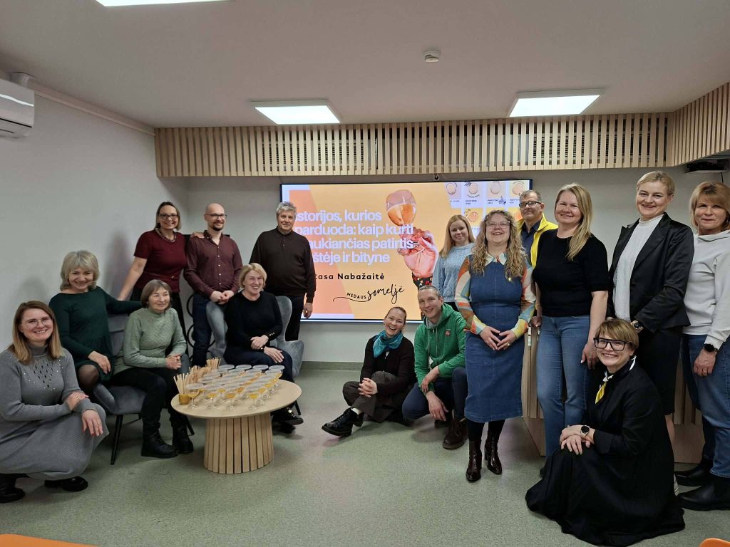 Group photo of participants at a workshop or event, gathered in a modern meeting space with a presentation screen displaying text in Lithuanian. Attendees are smiling and engaged, with drinks on a table in front of them, showcasing a collaborative atmosphere.