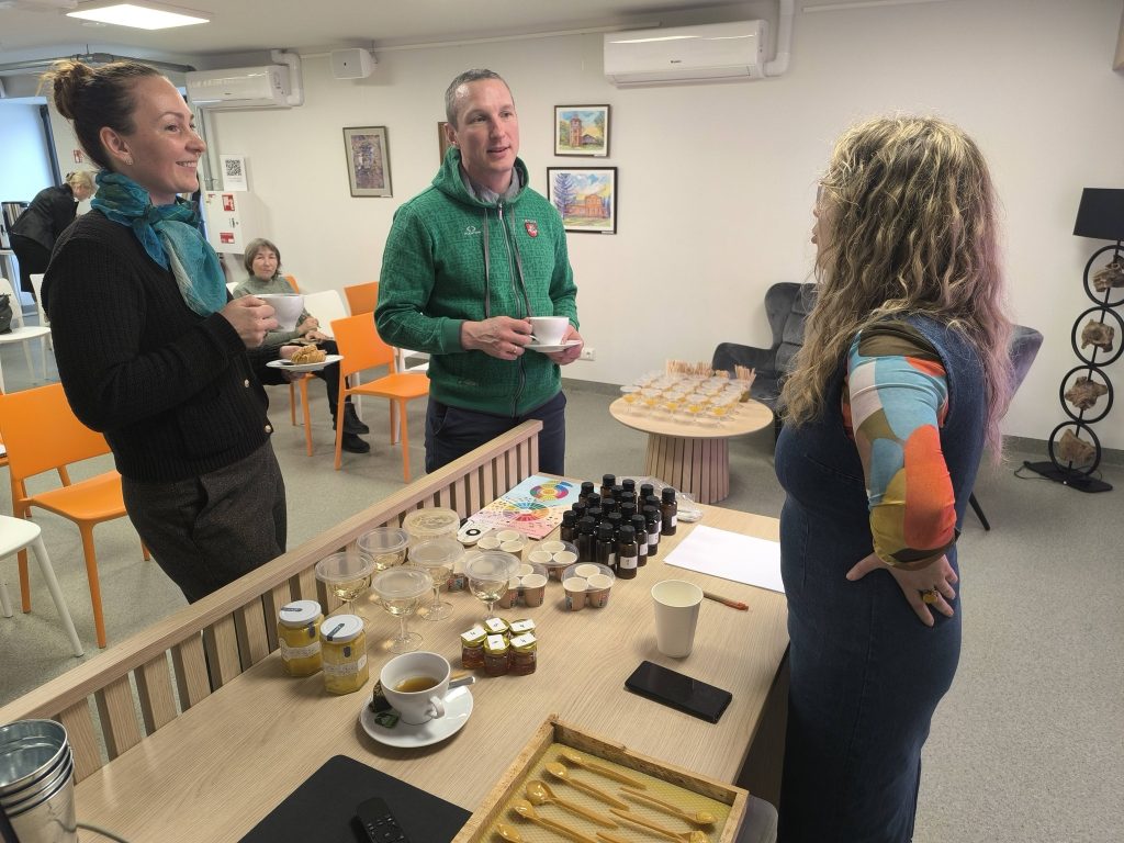 Networking event featuring three individuals engaged in conversation over tea and honey samples, with a display of jars and tasting cups on a table in a modern workspace. Bright orange chairs and a cozy atmosphere enhance the setting.