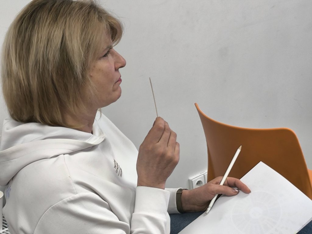 Woman with blonde hair holding a stick and a pencil, sitting in a classroom environment with an orange chair in the background. She appears to be deep in thought while looking at a sheet of paper.