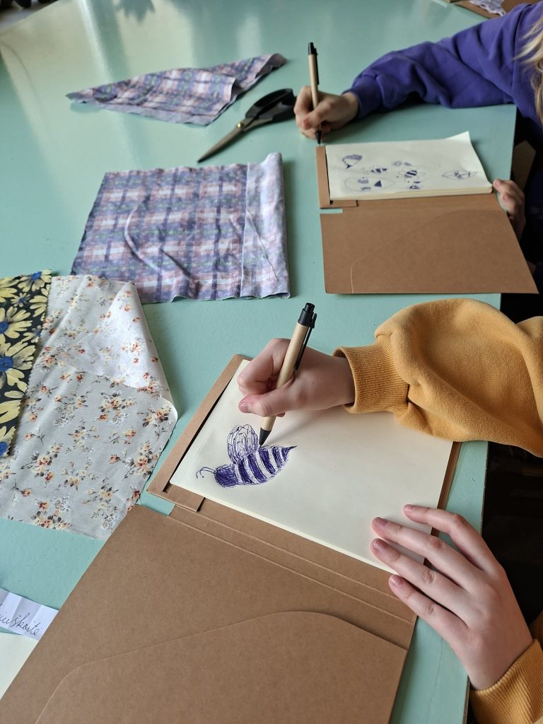Students engaged in a creative art session, sketching designs with pens on paper, surrounded by various colorful fabrics and scissors on a turquoise table.